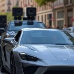 Cena de carro sendo perseguido por veículo tocando música em rua urbana espanhola.