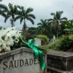 Buquê de flores brancas e laço verde em lápide de cemitério brasileiro, simbolizando luto por Lorena Batista, vítima de câncer nas vias biliares.
