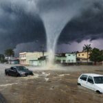 Cena de tromba d'água inundando rua em Goiânia, com enchente e chuva forte, sem pessoas.